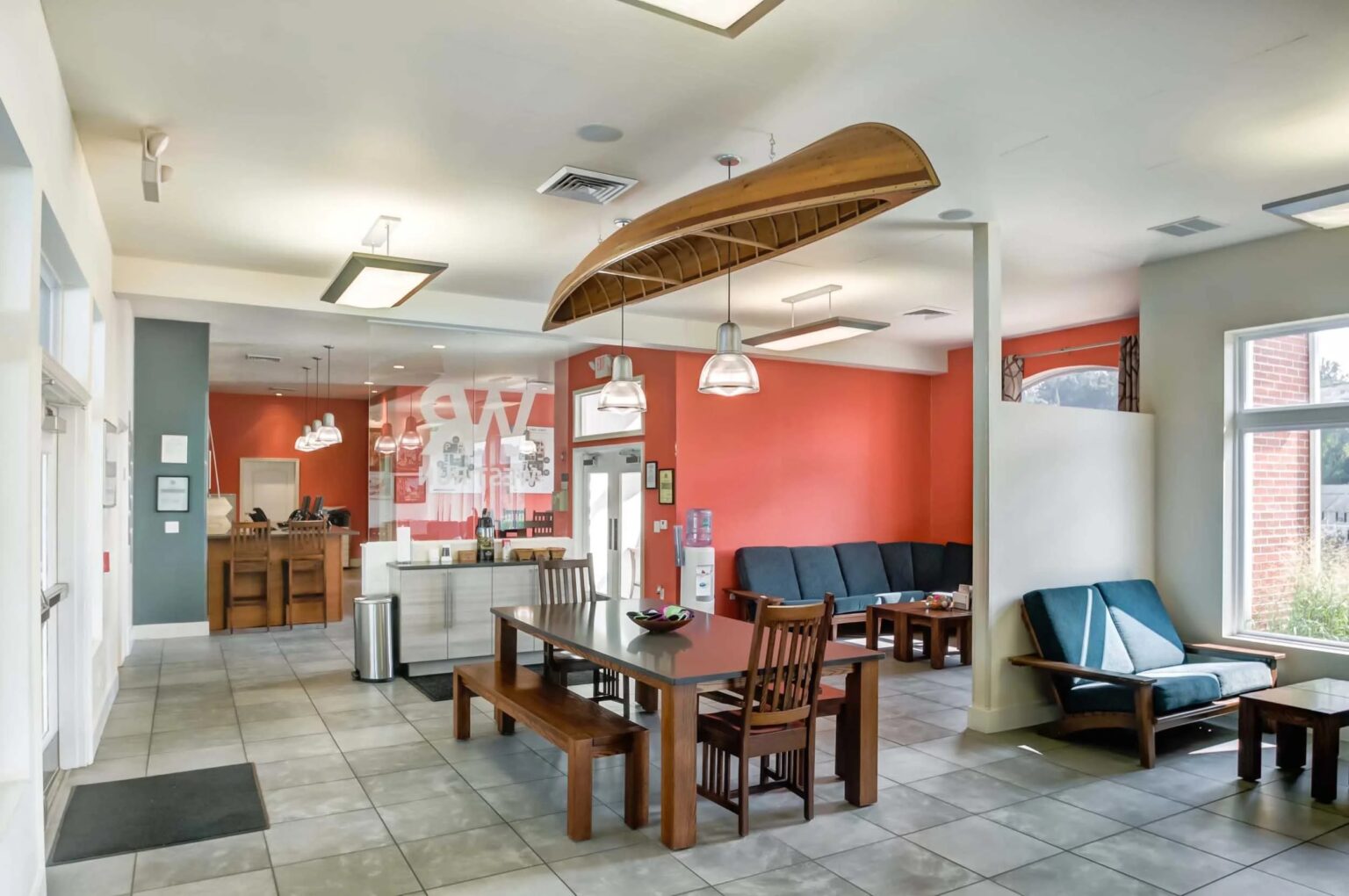 A modern lobby with tile flooring, wooden tables and chairs, a canoe hanging from the ceiling, a red accent wall, and a seating area with sofas near large windows.