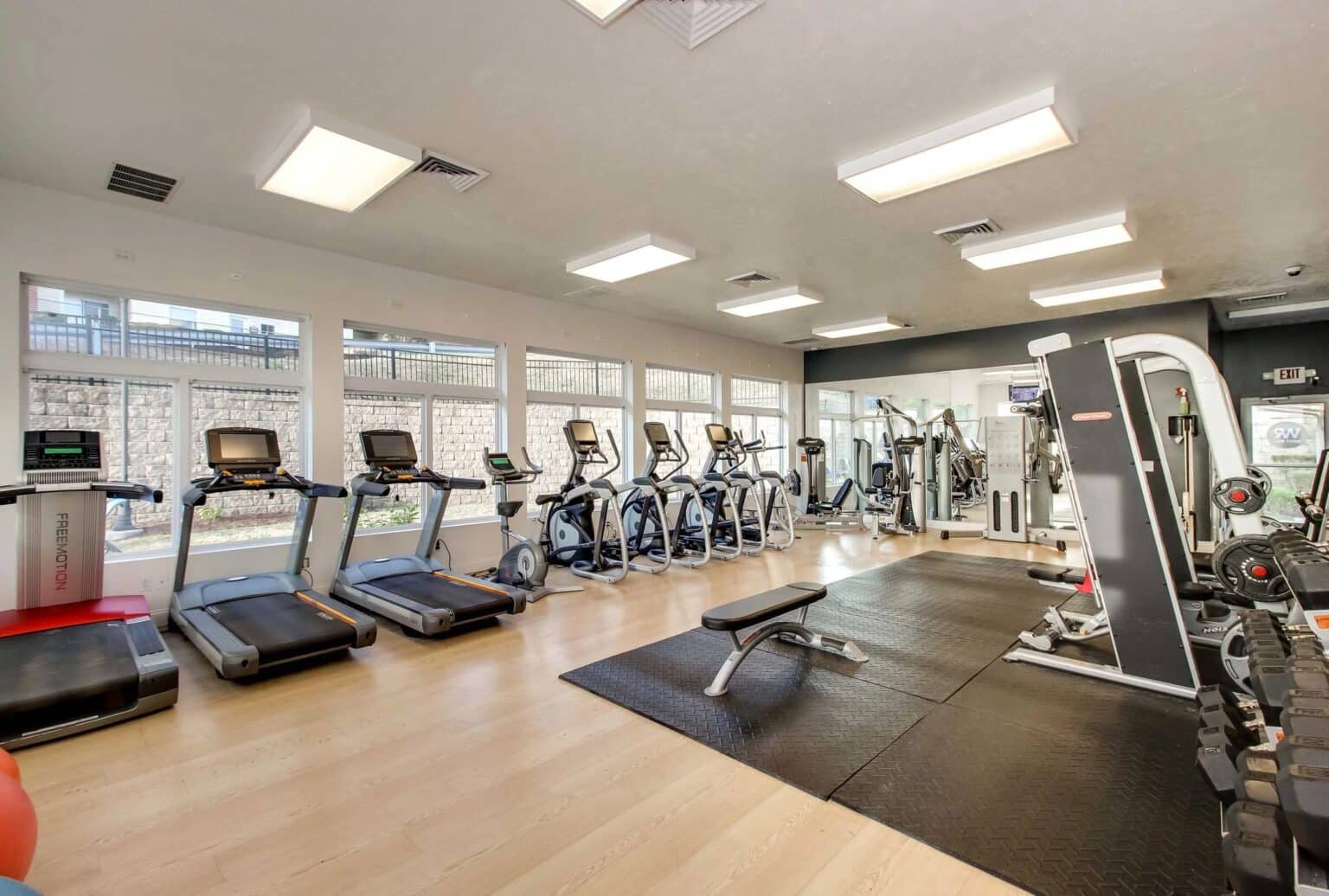 A well-lit gym with treadmills, elliptical machines, free weights, a bench, and strength training equipment on a wood and rubber floor.