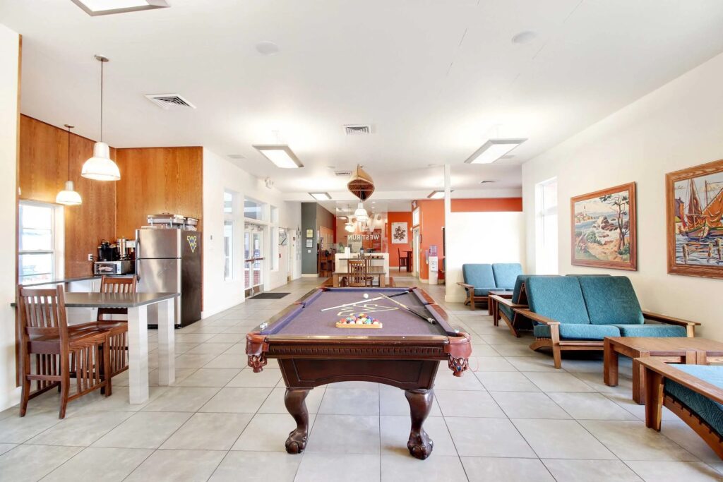 A spacious recreation room with a pool table in the center, lounge chairs, a dining table, a kitchenette, and artwork on the walls.