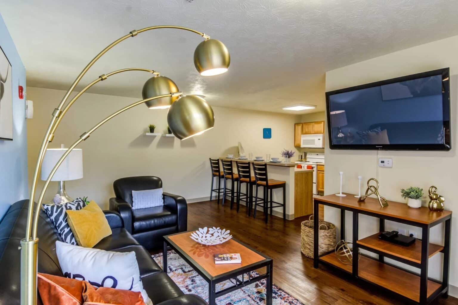 Modern living room with black leather furniture, a large gold floor lamp, mounted TV, and open kitchen with barstools; wood floors and neutral walls throughout.