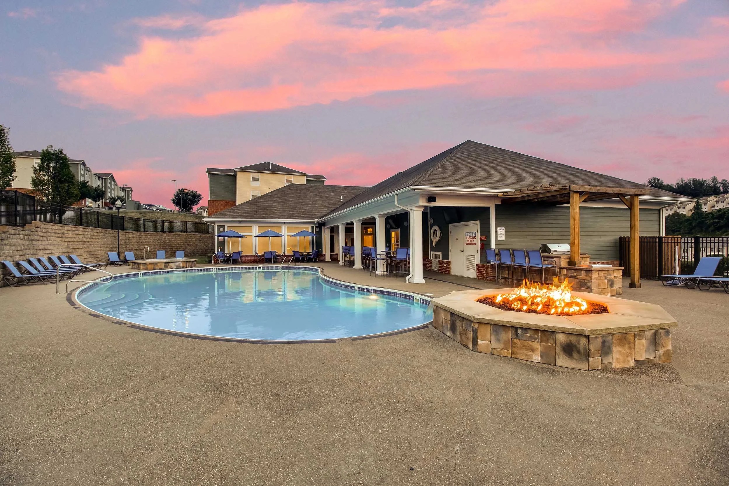 Outdoor swimming pool area with lounge chairs, a covered patio, and a lit fire pit in front of a building at sunset.