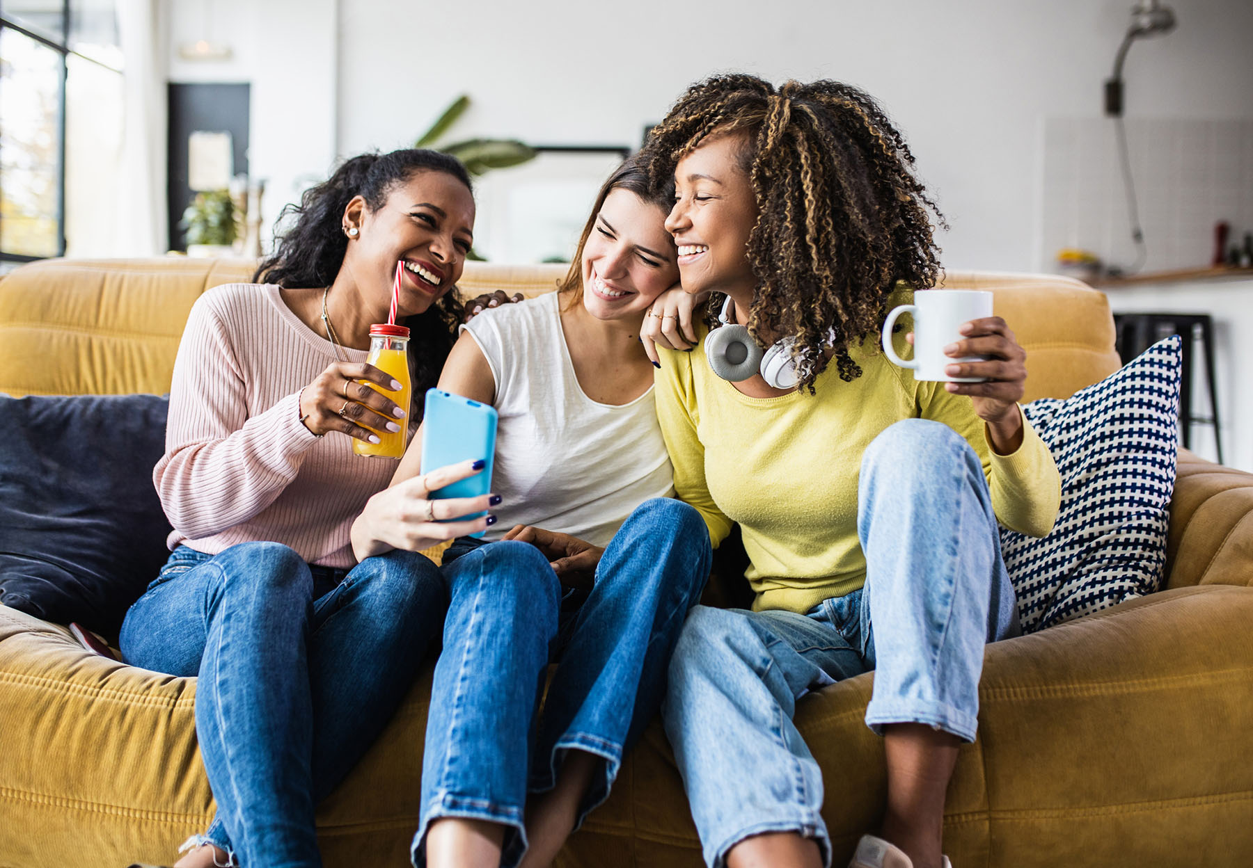 Three women sitting on a couch, smiling and taking a selfie together; one holds a drink, another a phone, and the third a mug with headphones around her neck.