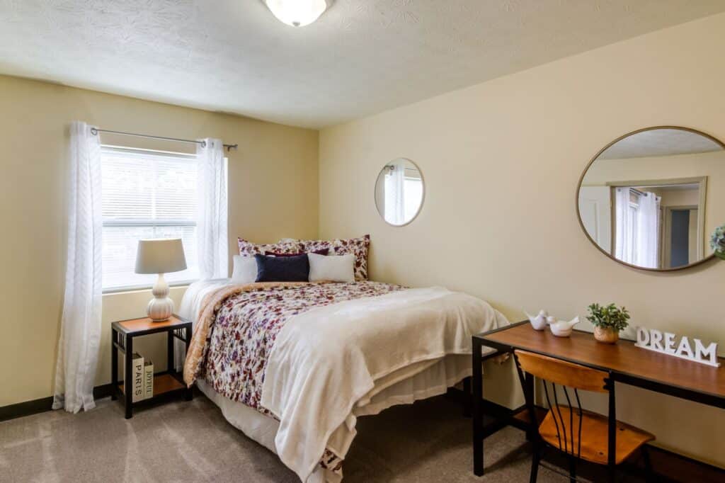 A neatly made bed with floral bedding, two round mirrors, a small desk with decor, and a window with white curtains create a cozy vibe in this softly lit Home - West Run bedroom.