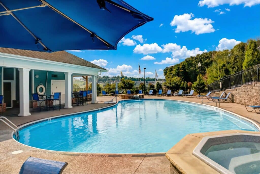 Outdoor swimming pool with adjacent hot tub, lounge chairs, and a covered seating area under a large blue umbrella await you at Home - West Run, all surrounded by trees and a blue sky with clouds.